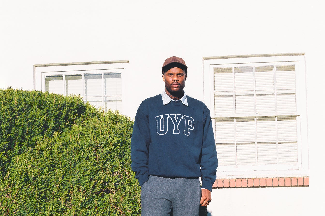 Man wearing a navy blue sweater with 'UYP' text, standing in front of a white house with greenery.